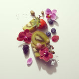 A styled flat-lay of fresh fruits and flowers on a clean white background, including sliced kiwi, a strawberry, blueberries, a blackberry, pink orchids, and small petals with water droplets.